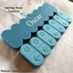 A customized 3D-printed weekly pill box set in medium size featuring two-tone blue colors. Each day’s compartment has sliding lids with engraved sun and moon symbols. A large nameplate behind the set shows an example name “Oscar” engraved to demonstrate the personalized name option. The image highlights the Medium + Name + Two Colors variation.