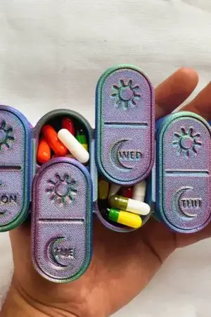 USACUTTIE iridescent weekly pill box in rainbow color-shift finish, seven daily compartments with sun and moon AM PM markings on the sliding lids