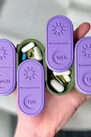 Hand holding the USACUTTIE weekly pill organizer with MON, TUE, WED and THU compartments open showing pills inside, sized to fit in one hand