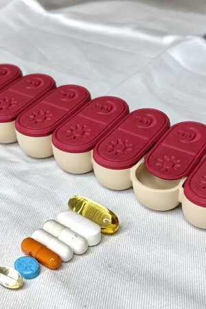 USACUTTIE fall pill organizer with cranberry red lids and cream compartments, capsules and tablets lined up in front on a white sheet