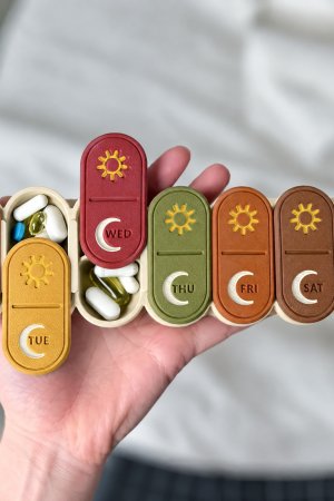 USACUTTIE 7 day AM PM pill organizer in a fall special mix held in one hand: Pumpkin, Mustard, Cranberry, Olive, Rust, Cinnamon and Caramel lids with gold sun and ivory moon icons on a cream box